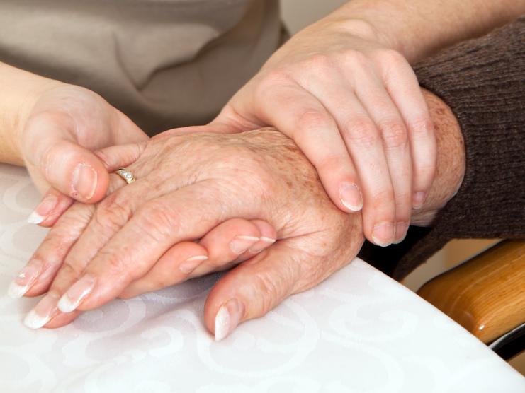 Zwei Hände, eine ältere und eine jüngere, liegen auf einem Tisch, wobei die jüngere Hand die ältere sanft umschließt.