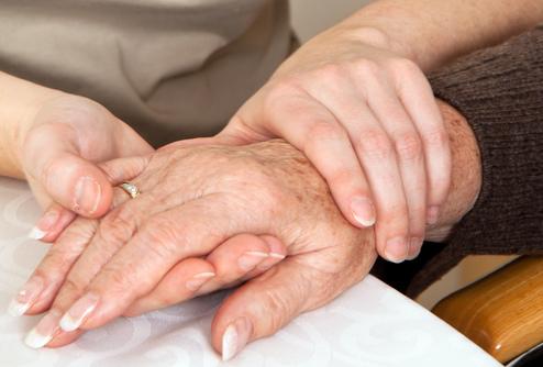 Zwei Hände, eine ältere und eine jüngere, liegen auf einem Tisch, wobei die jüngere Hand die ältere sanft umschließt.
